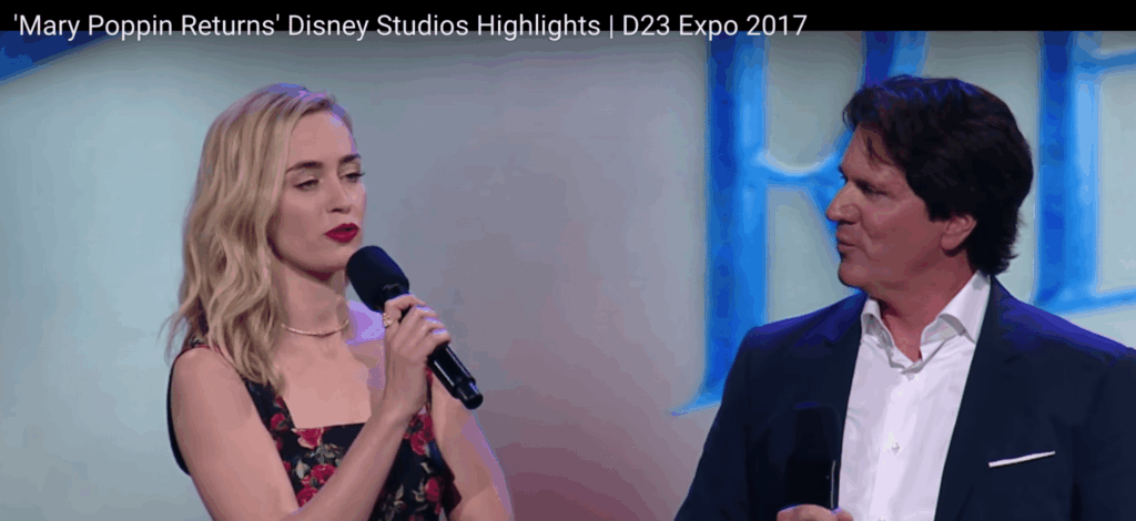A blonde woman speaks into a microphone beside a man in a dark suit on a stage for Mary Poppins, with blue-lit panels in the background.
