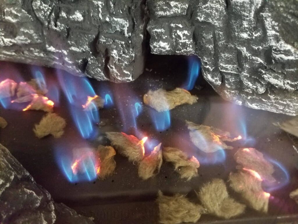 Blue and orange flames burn among tan faux logs inside a dark faux-stone fireplace.