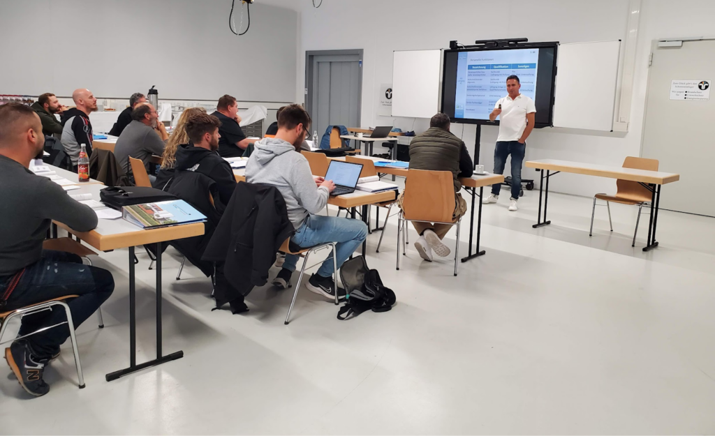 A man presents at the front of a classroom to seated adults using a large screen, while attendees take notes with laptops and tools at wooden desks.
