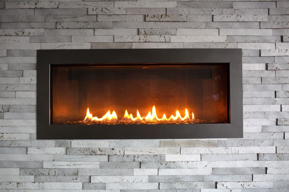Modern horizontal gas fireplace insert built into a textured stone wall, showing the clean installation and the need for periodic cleaning and maintenance of the burners and glass panel.