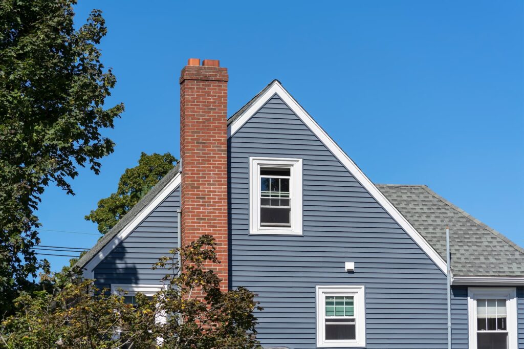 Blue-gray house with a brick chimney and white-trimmed windows under a clear blue sky, illustrating how to tell if damper is open or closed, how to open damper on fireplace, and how to open fireplace flue.