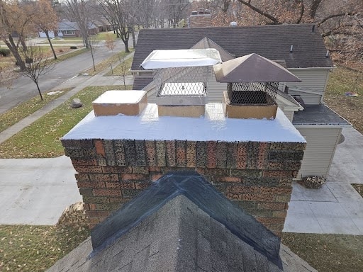 A brick chimney with three metal flue caps and a shingled roof in the foreground, viewed from above with a house, driveway, and trees behind it.