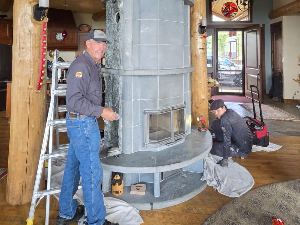 Two workers install a large gray tiled fireplace in a wood-paneled room with a ladder, tools, and an open doorway in the background.