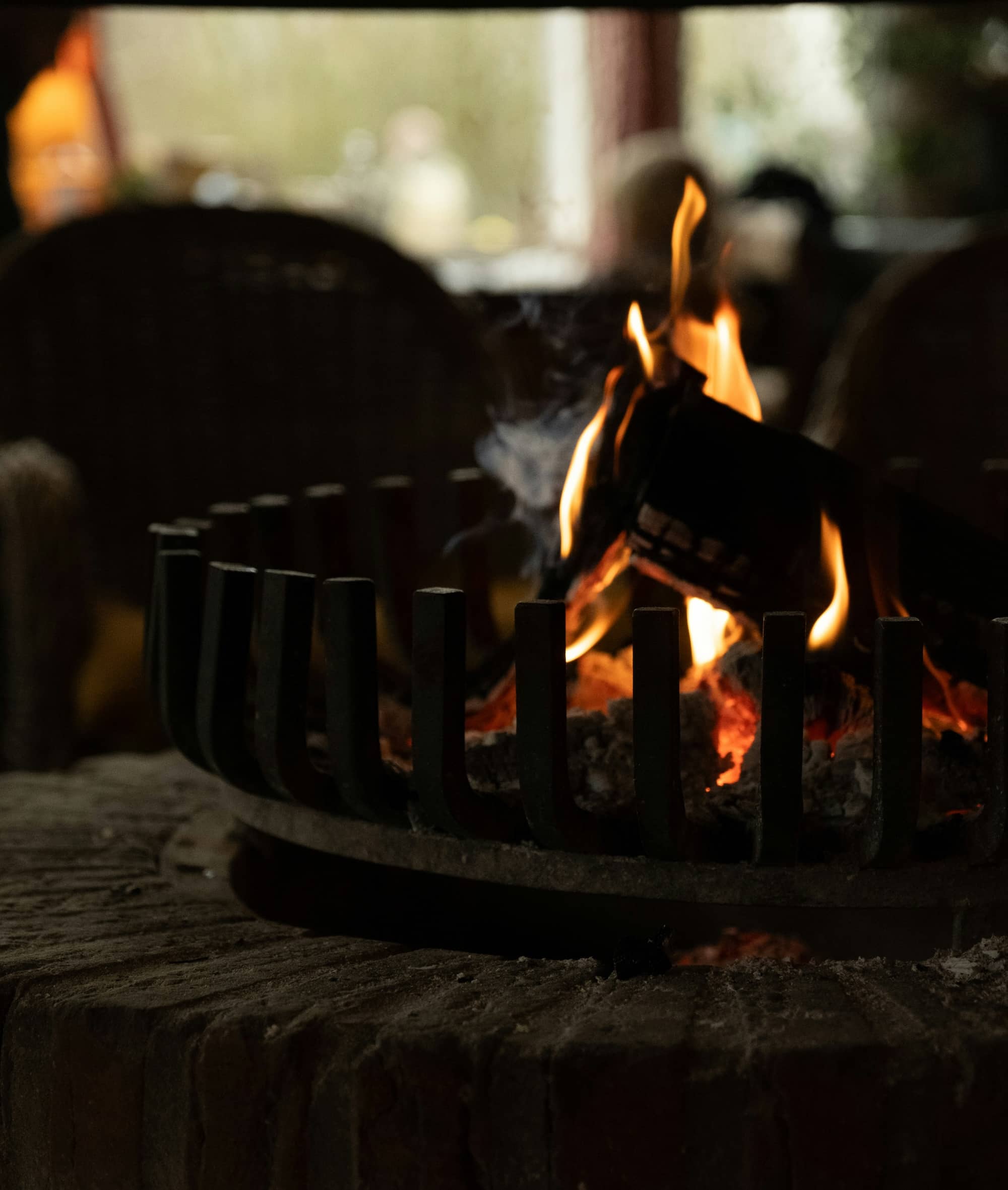 A burning log sits in a circular metal fire grate on a stone hearth, with a dimly lit room in the background.