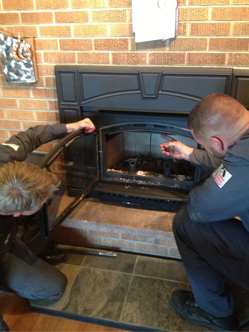 Two workers inspect a black fireplace insert with the glass door open, crouched in front of a brick wall and tiled hearth.