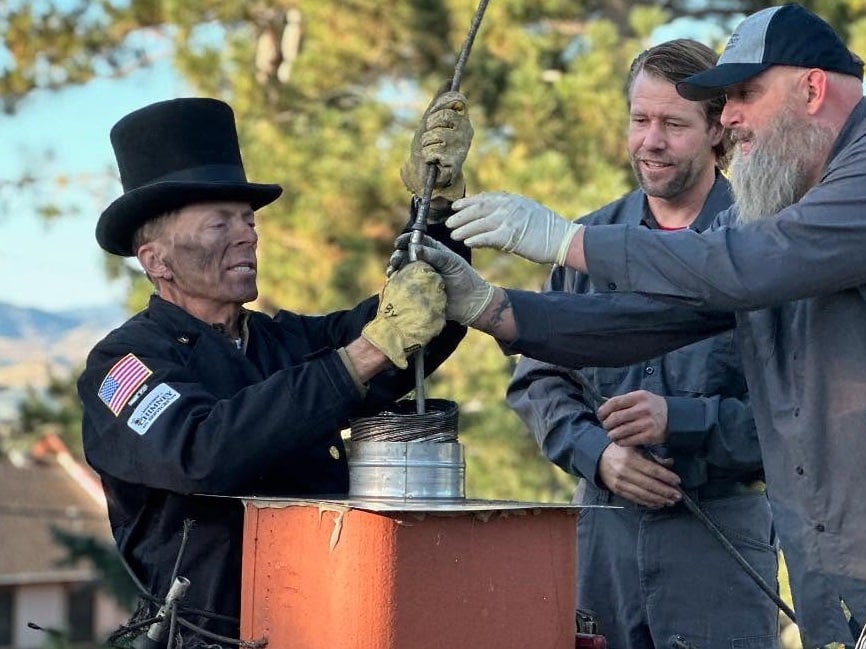 Three men work together to lift a metal chimney pipe from an orange structure outdoors, with trees and a house in the background.