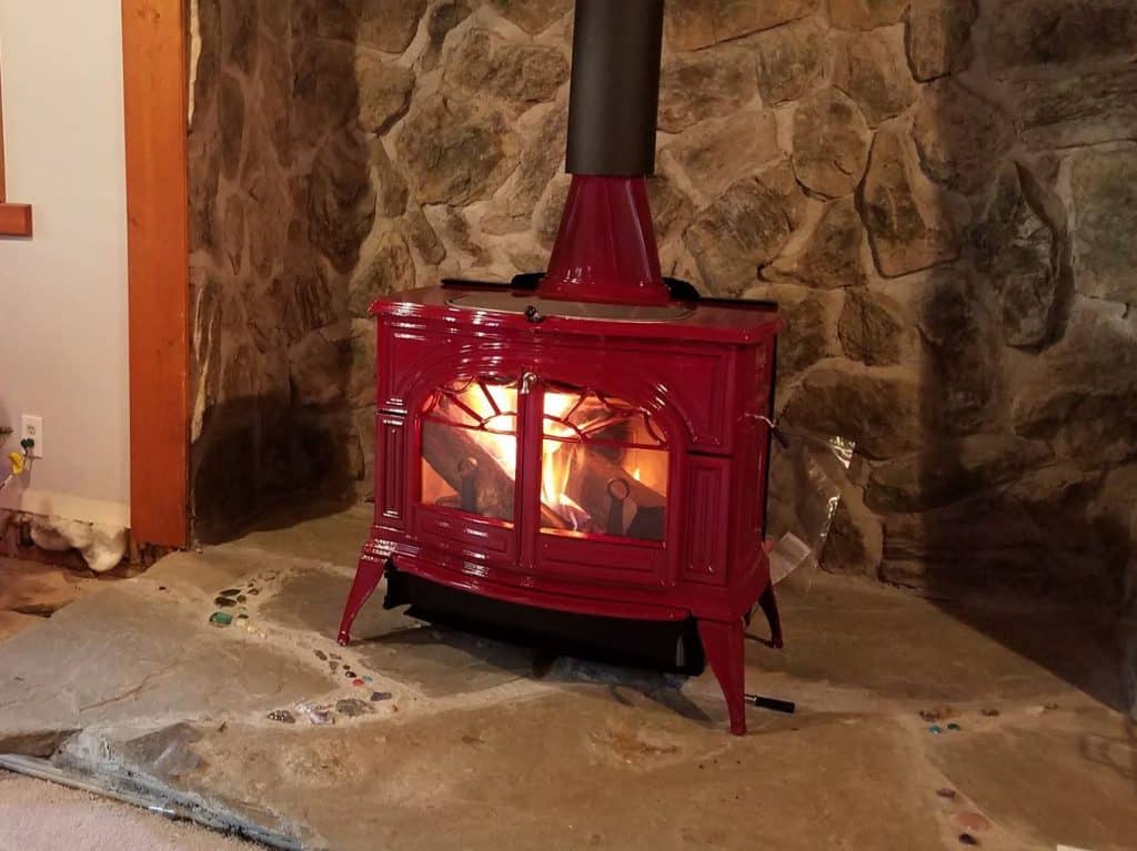 A red wood stove with a black chimney pipe sits on a stone hearth against a rough stone wall.