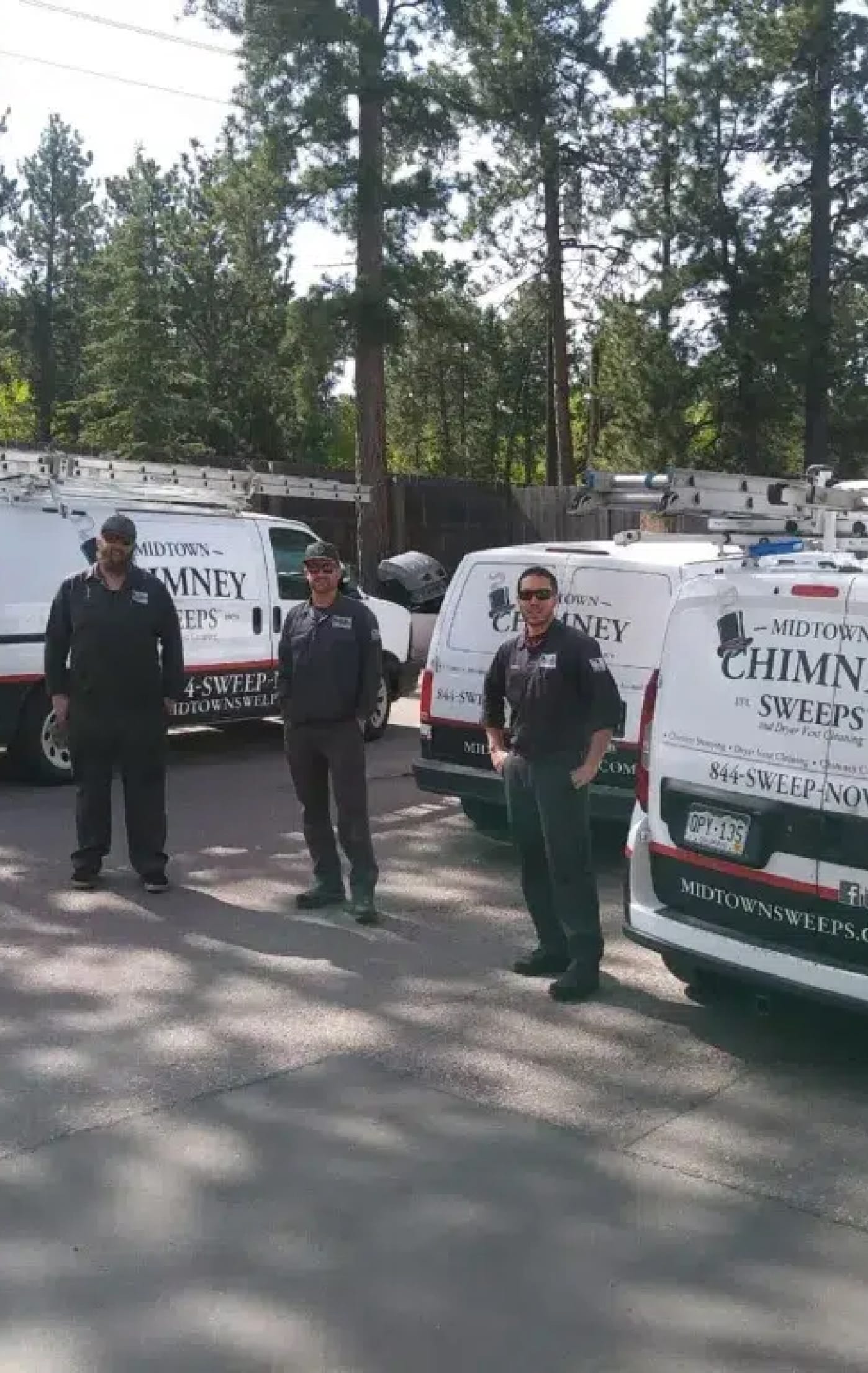 Three chimney sweeps stand beside white service vans parked along a tree-lined road.
