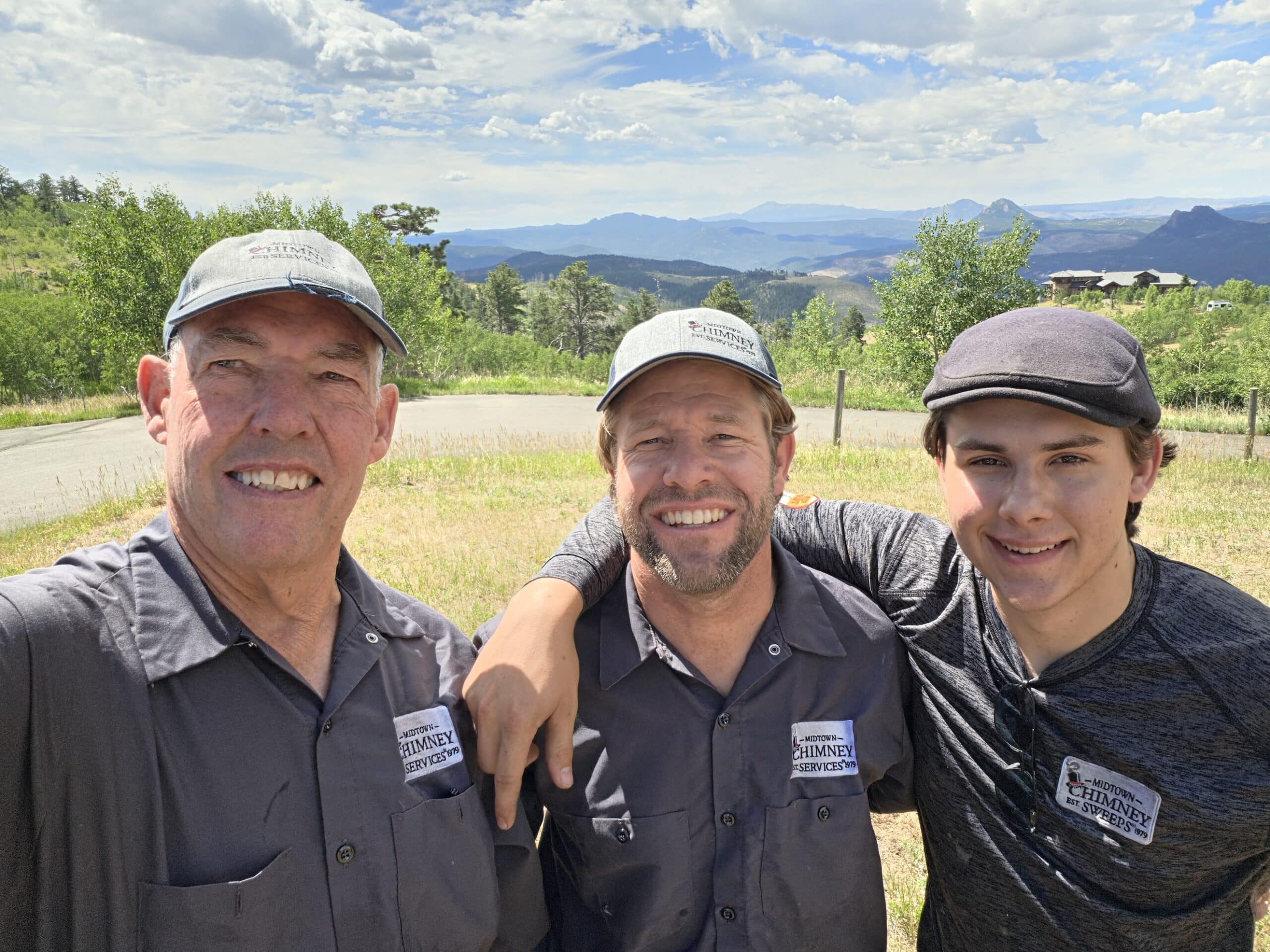Three men wearing caps and dark work shirts stand together outdoors with mountains, trees, and a winding road in the background.