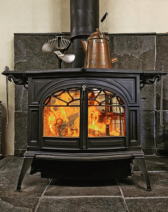 A black wood-burning stove with visible flames behind arched glass doors, topped by a copper kettle and set against a dark stone hearth.