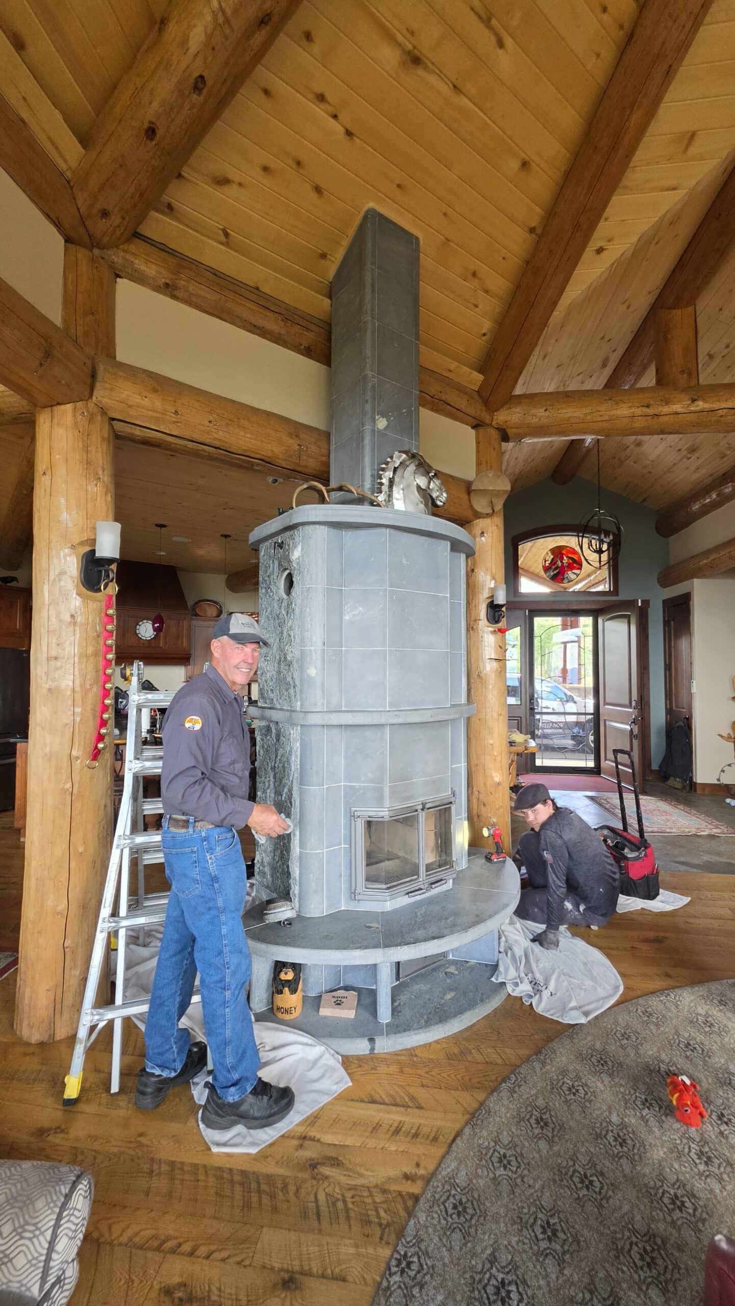 A man stands beside a tall gray wood stove in a log cabin interior with exposed beams, while another man kneels on the floor nearby.