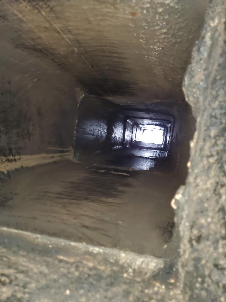 A dirty rectangular ventilation duct extends into a bright opening, with grime coating the metal interior.
