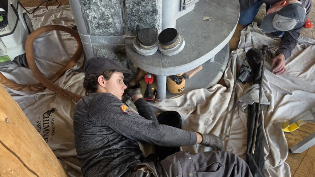 A worker in gray clothes installs a black pipe beneath a round metal structure while another worker crouches beside supplies on a protective tarp.