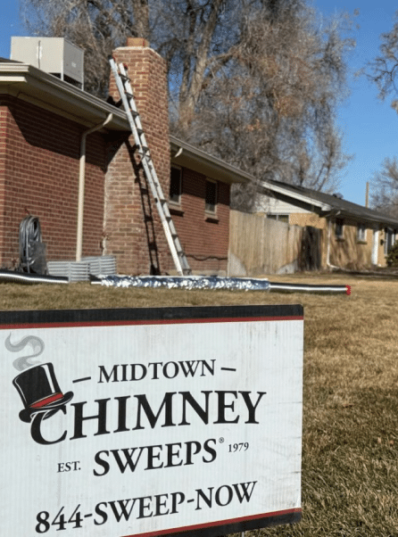 A Midtown Chimney Sweeps sign stands in front of a brick house with a ladder leaning against the chimney and a hose on the lawn.