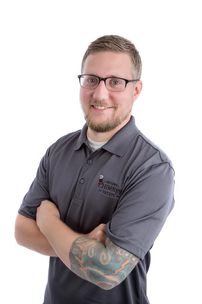 A man with glasses and a tattooed forearm stands with folded arms, wearing a gray polo shirt against a white background.