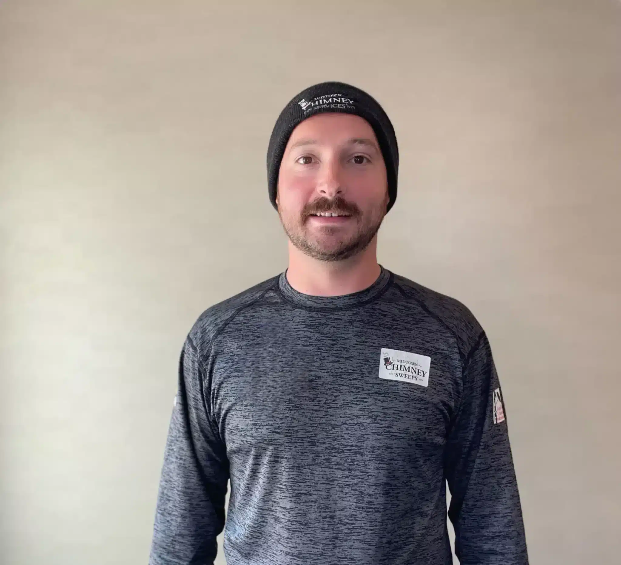 A man wearing a black beanie and gray long-sleeve shirt with a Chimney label stands against a plain beige wall.