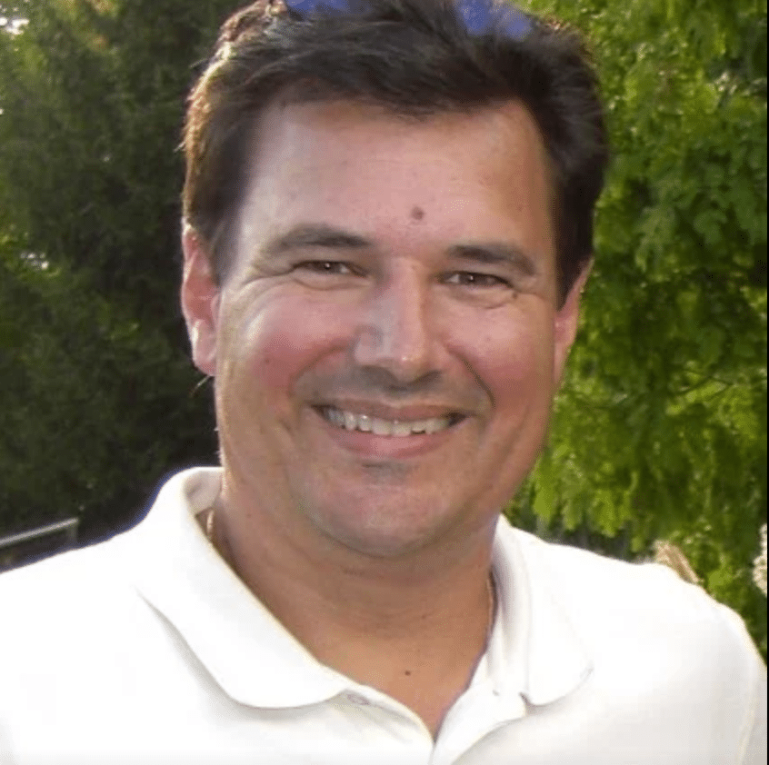 A smiling man in a white polo shirt, photographed outdoors with green trees in the background.