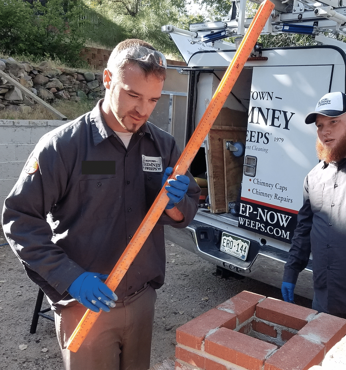 A worker in a gray uniform and blue gloves measures an orange rod beside a brick chimney, with a chimney service truck and another worker in the background.
