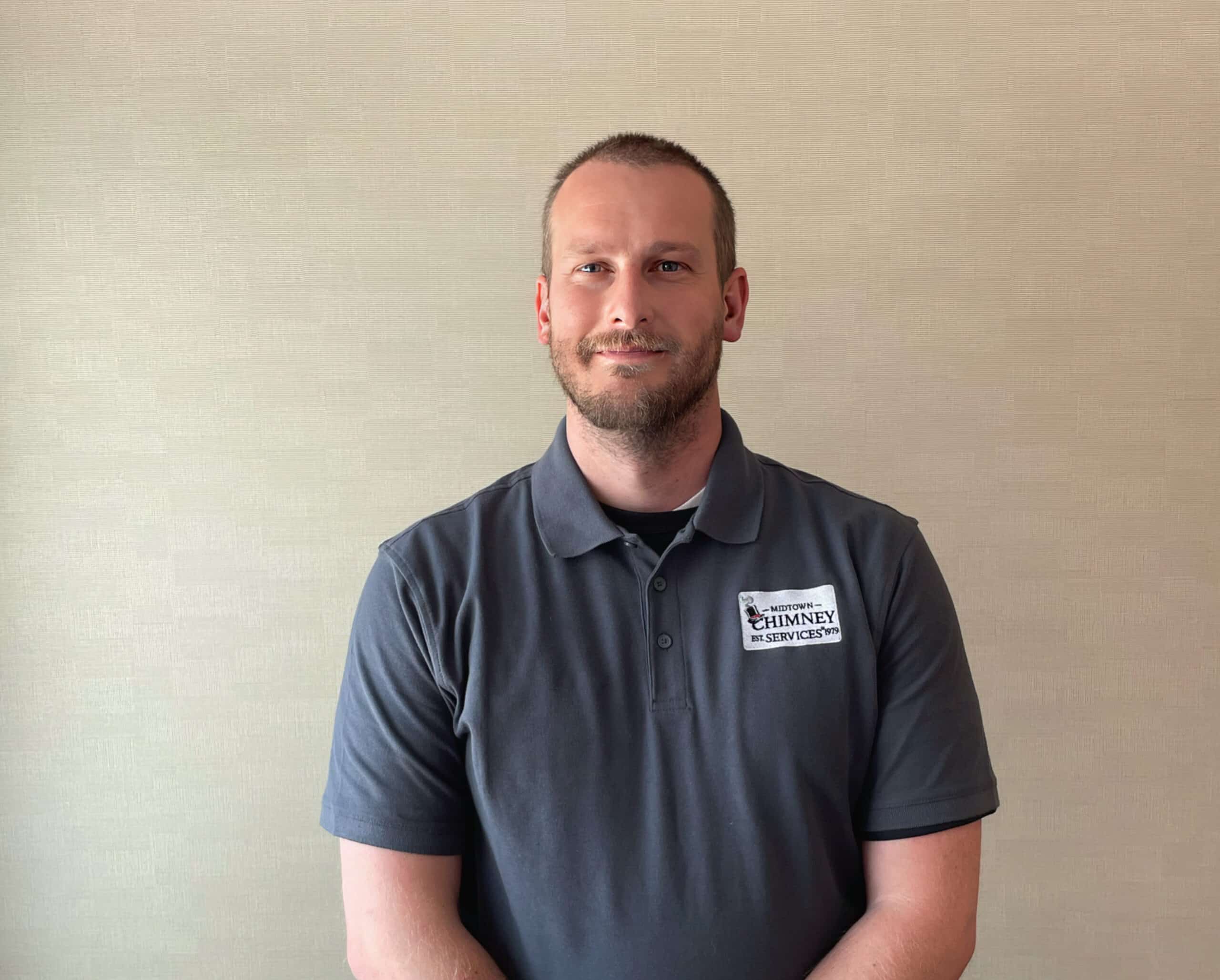A man in a dark gray polo shirt with a Chimney Services logo stands against a plain beige wall.