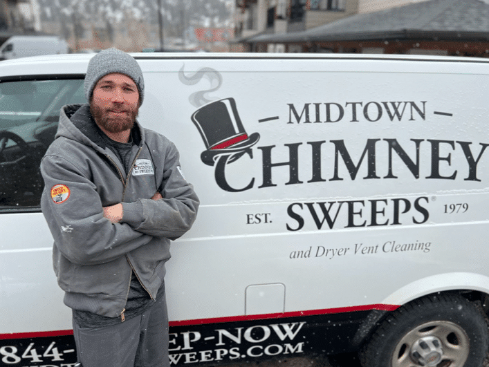 A man in a gray beanie and work jacket stands beside a Midtown Chimney Sweeps van with the company logo and contact text on the side.