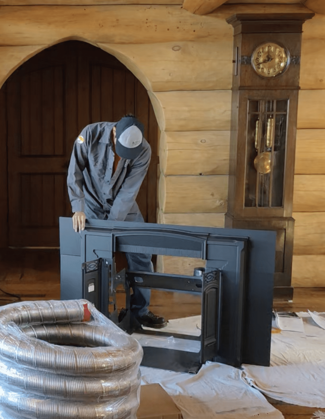A worker in a gray jacket and cap installs a large black television in a log cabin, with tools, cords, and coiled ducting on a covered floor.