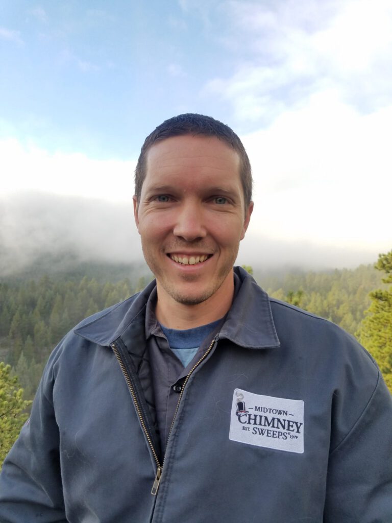 A man in a blue work jacket with a Mid-Down Chimney Sweeps logo stands outdoors, with trees, fog, and a cloudy sky in the background.
