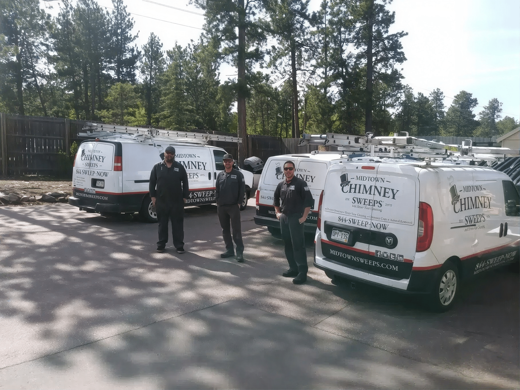 Three workers stand beside white chimney cleaning service vans parked on a paved lot with trees and a wooden fence in the background.