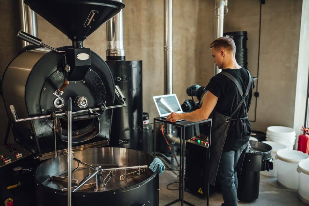 A man operates a coffee roaster and laptop in a roasting room with stainless steel equipment and white buckets.