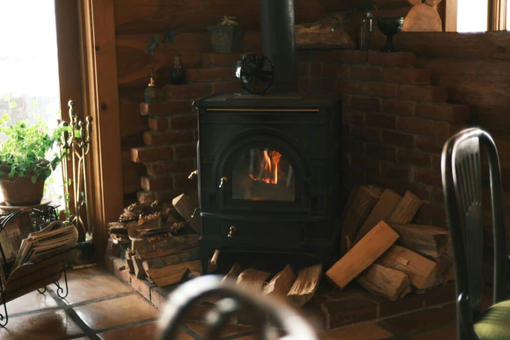 A black wood-burning stove with a fire burning inside sits against a brick hearth, with stacked firewood and a potted plant by the window.