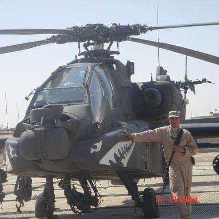 A military helicopter with shark-mouth nose art sits on a tarmac beside a man in a tan flight suit.