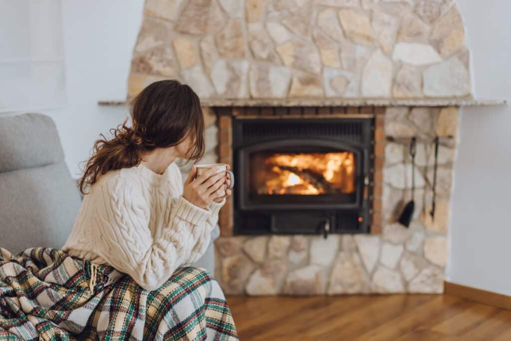 A woman in a cable-knit sweater sits by a stone fireplace holding a mug, with a lit wood stove and plaid blanket in the living room.