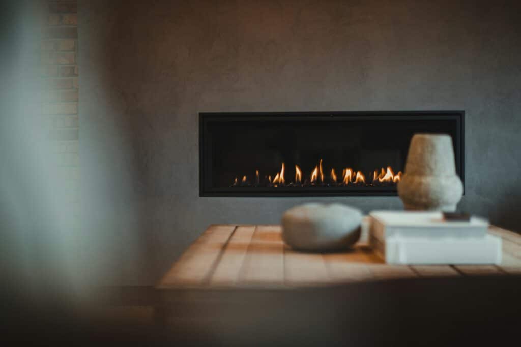 A modern wall-mounted fireplace with small flames, set behind a wooden table holding a stone bowl and stacked books.