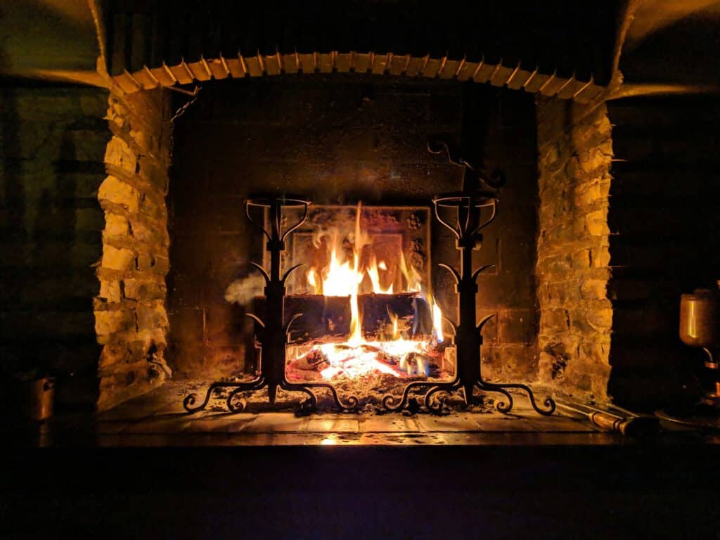 A lit fireplace with flames burning logs behind black andiron tools, framed by a stone hearth and dark interior walls.