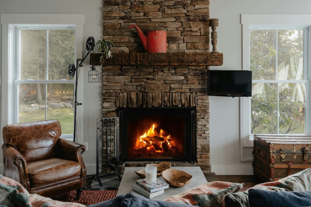 A leather armchair sits beside a stone fireplace with a burning fire, with windows on both sides and a coffee table in the foreground.