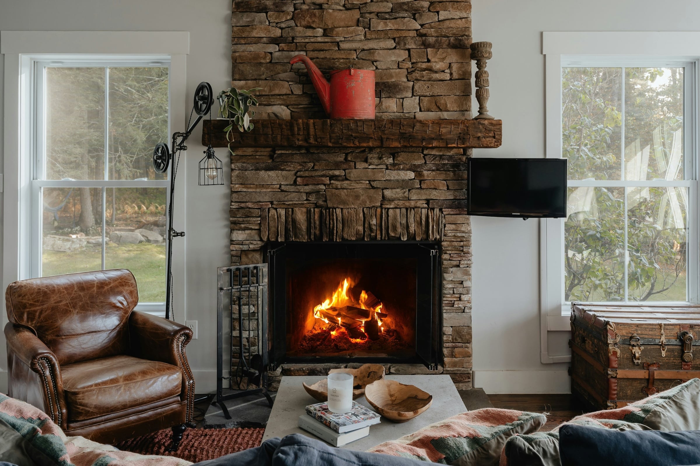 A leather armchair sits beside a stone fireplace with a burning fire, with windows on both sides and a coffee table in the foreground.