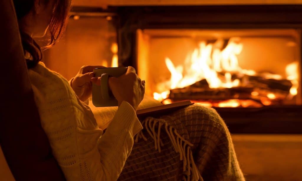 A person in a light sweater holds a mug beside a glowing fireplace with visible flames.