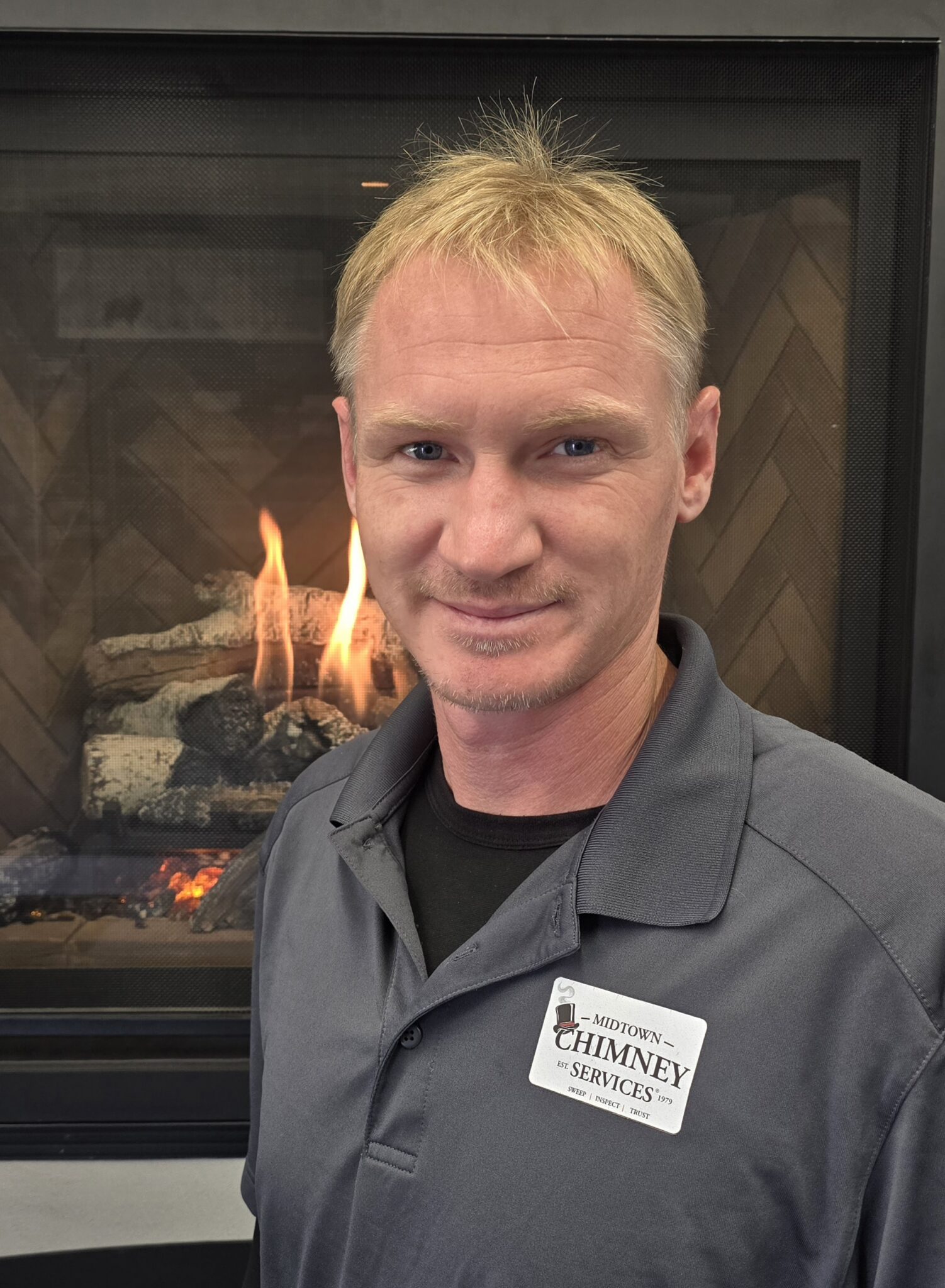 A blond man wearing a gray Chimney Services polo shirt stands in front of a lit fireplace with glass doors.