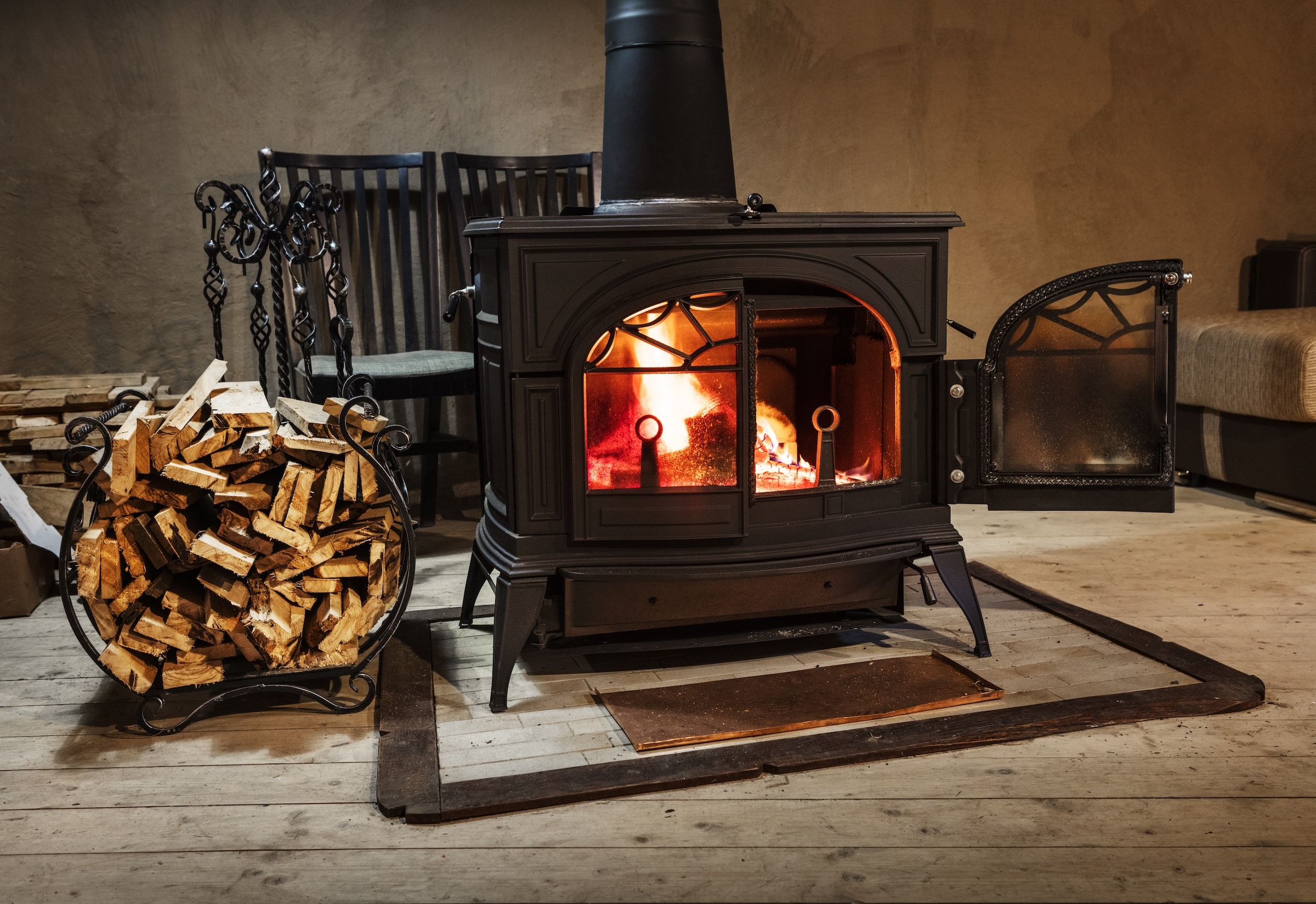 A black wood-burning stove with a visible fire inside stands on a wooden floor, with a log basket and stacked chairs beside it.