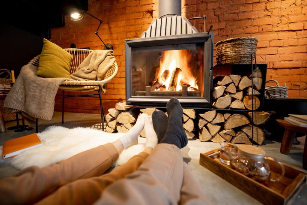 A fireplace with burning logs sits in a brick-walled room beside a cushioned wicker chair, with two people’s feet resting on a white rug in the foreground.