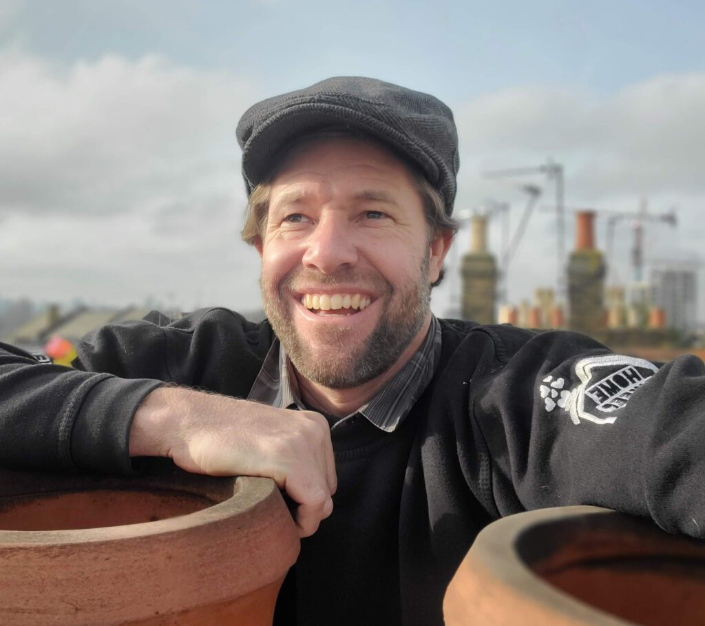 A smiling man wearing a flat cap and black sweater leans on terracotta pots, with industrial structures and a cloudy sky in the background.