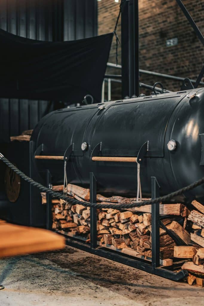 A large black barrel smoker filled with stacked firewood, set against a brick wall and dark draping in an industrial space.