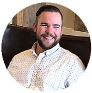 A smiling man with short dark hair and a beard sits in a chair, wearing a white patterned button-up shirt.