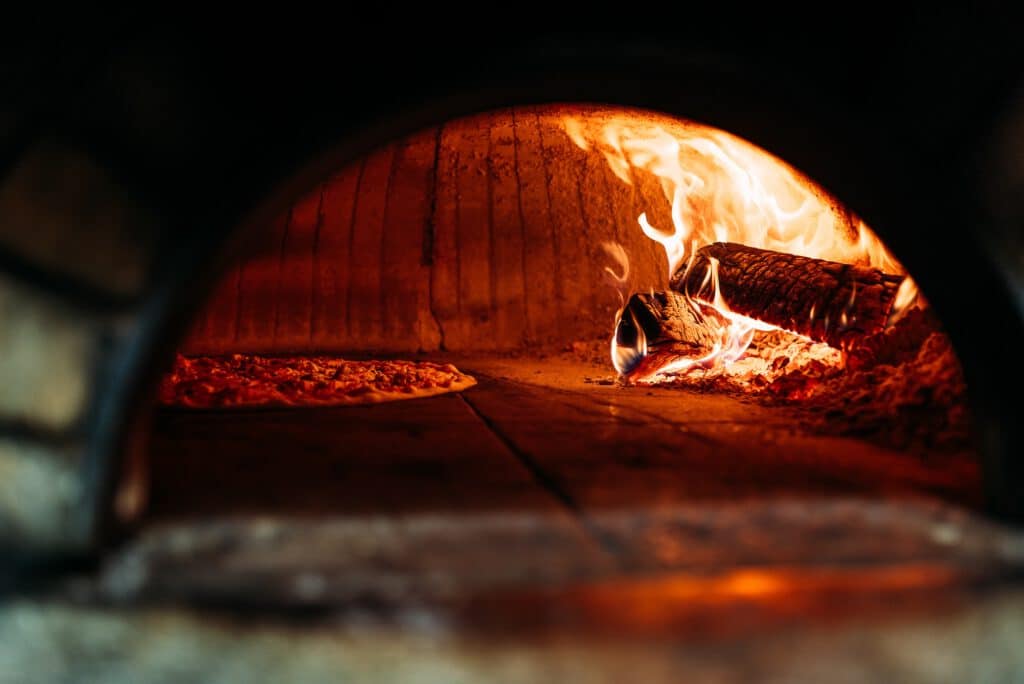 A pizza bakes inside a wood-fired oven, with flames along the right side and a pizza on the left.